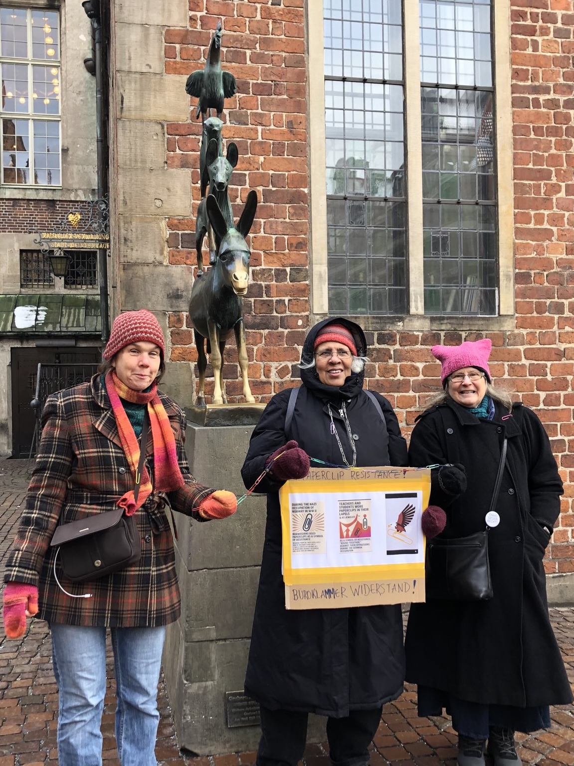 A group of three women stands in front of the Bremen Town Musicians monument. One woman is holding a sign displaying information about the paperclip campaign that symbolizes resistance and solidarity.